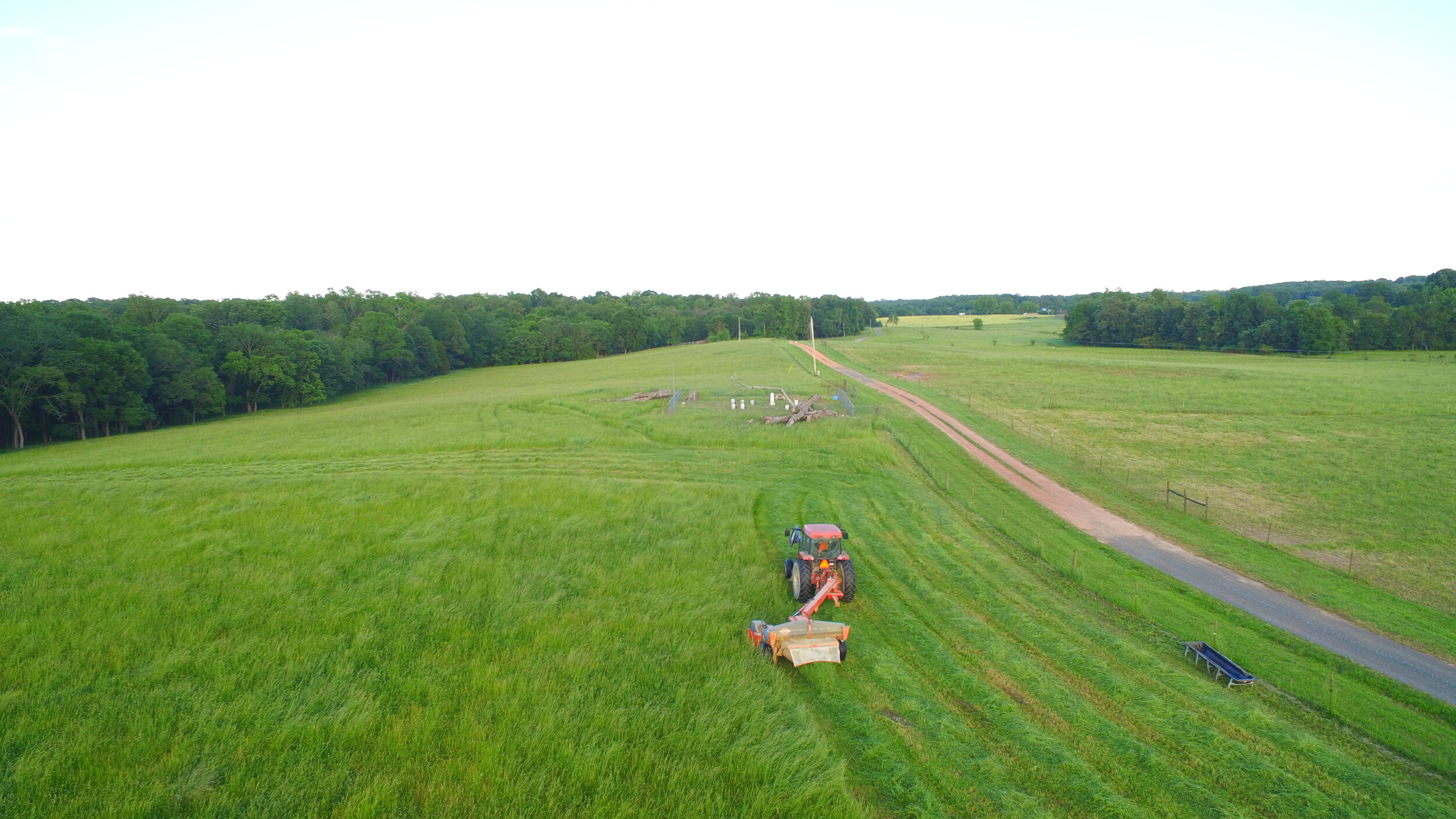 Aerial view of a green pasture with a tractor mowing grass, surrounded by trees and a dirt road, showcasing rural farming landscape.