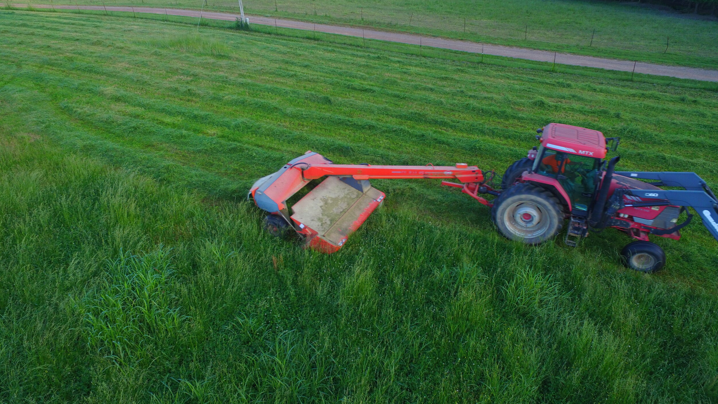 Jeremy+Brooks+-+DJI_0010 Aerial view of a red tractor using a mower to cut grass in a lush green field, with a dirt road in the background.