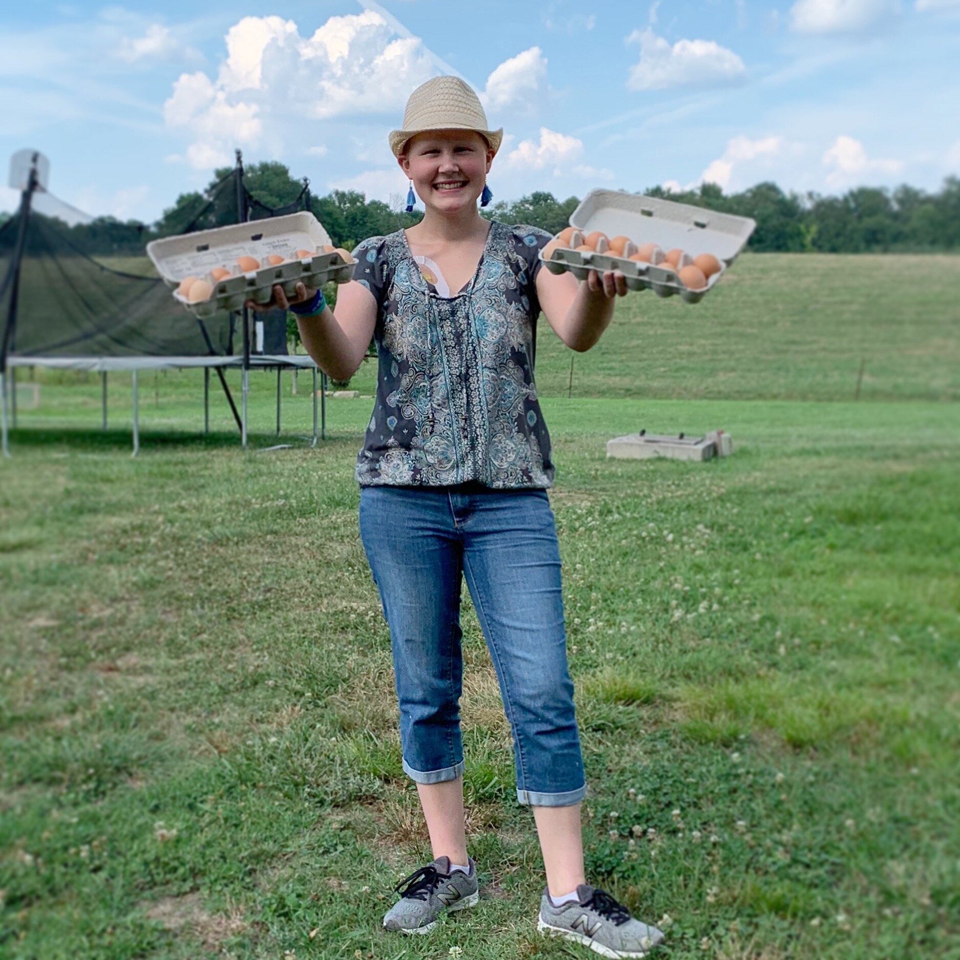 Jeremy+Brooks+-+Liz+&+Eggs Smiling woman holding two cartons of eggs outdoors on a sunny day, wearing a straw hat and casual attire, with a grassy field and trampoline in the background.