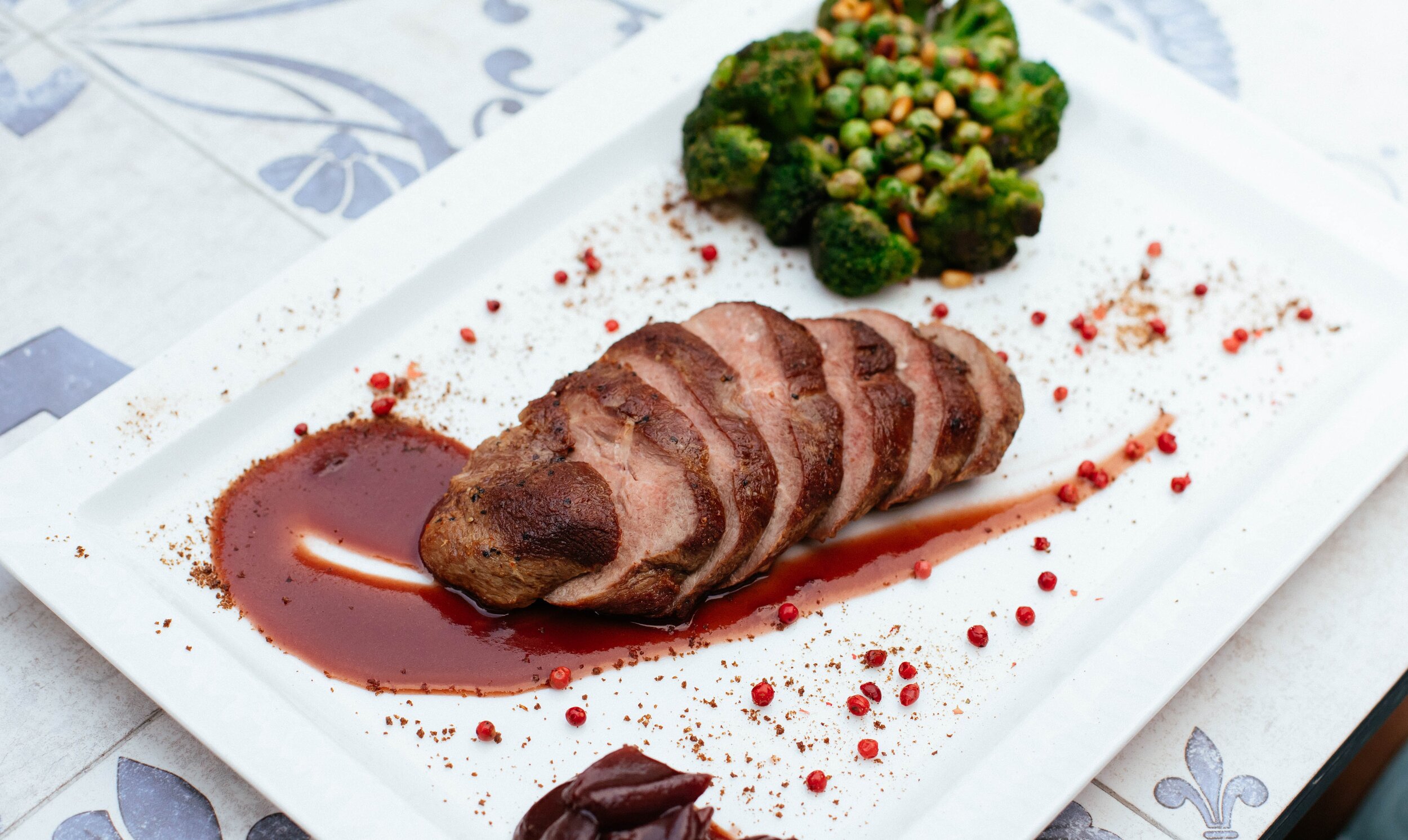 Grilled and sliced duck breast served with a rich red sauce, accompanied by steamed broccoli and peas, garnished with pink peppercorns on a white rectangular plate.
