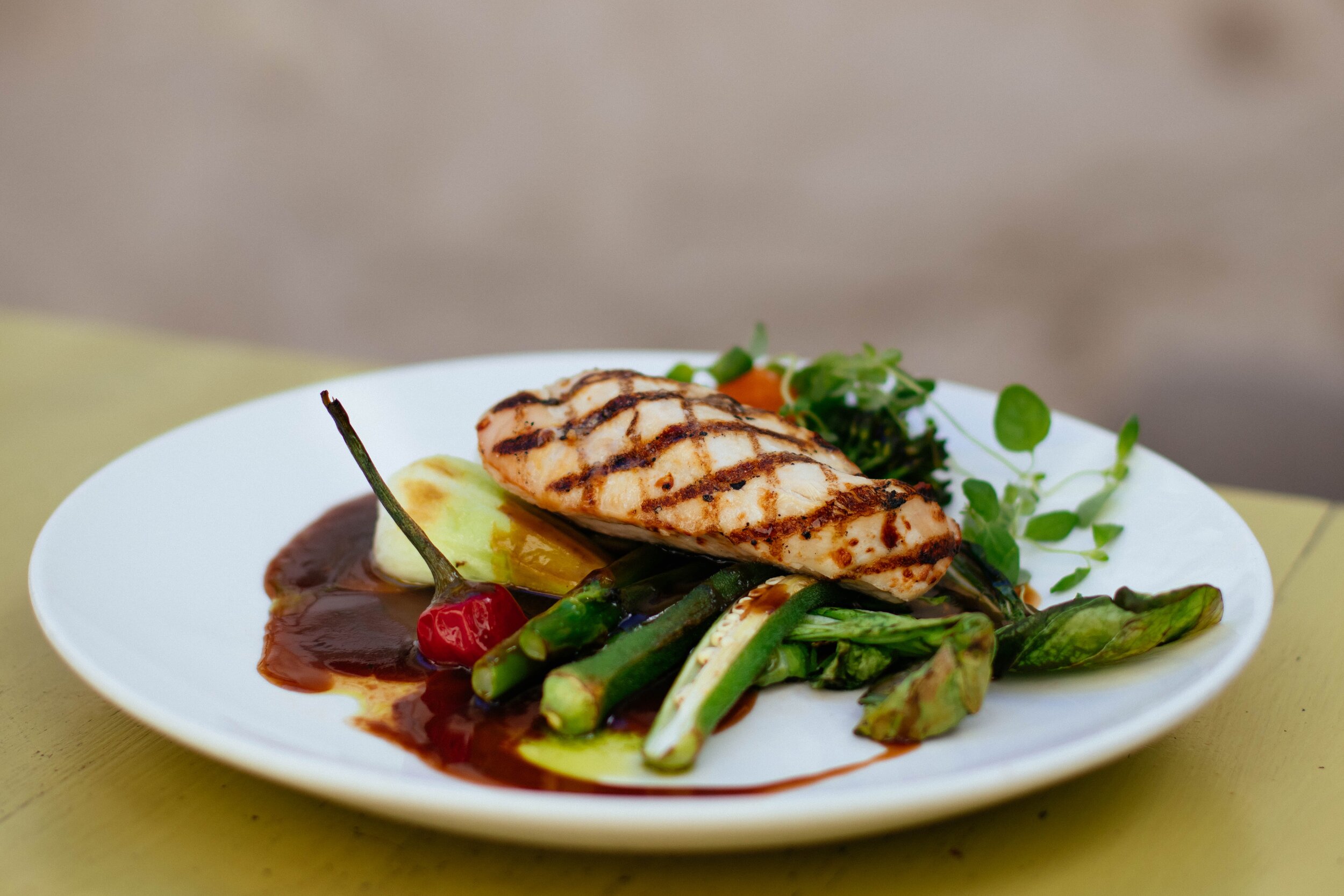 Grilled salmon fillet served on a white plate, accompanied by a colorful mix of sautéed vegetables including asparagus, cherry tomatoes, and greens, drizzled with a rich sauce.
