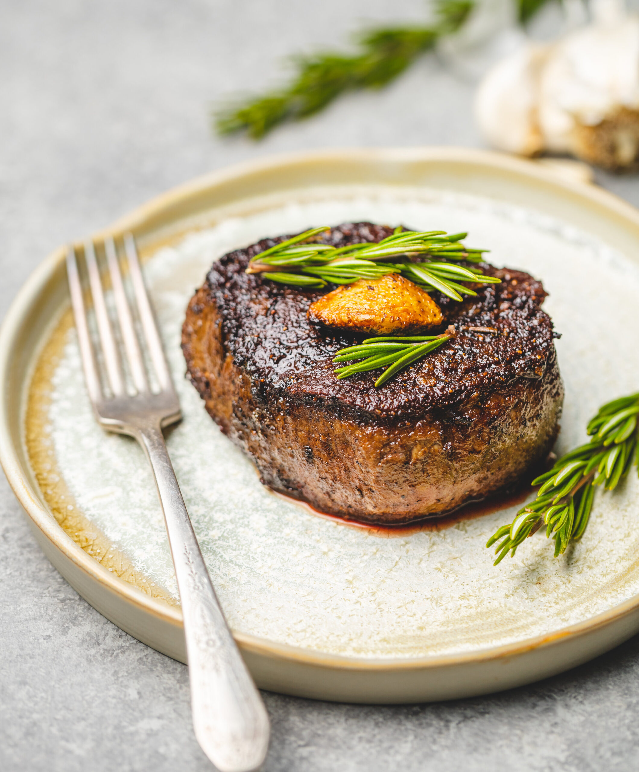 Juicy grilled filet mignon topped with fresh rosemary and garlic, served on a rustic plate with a fork, showcasing a gourmet dining experience.